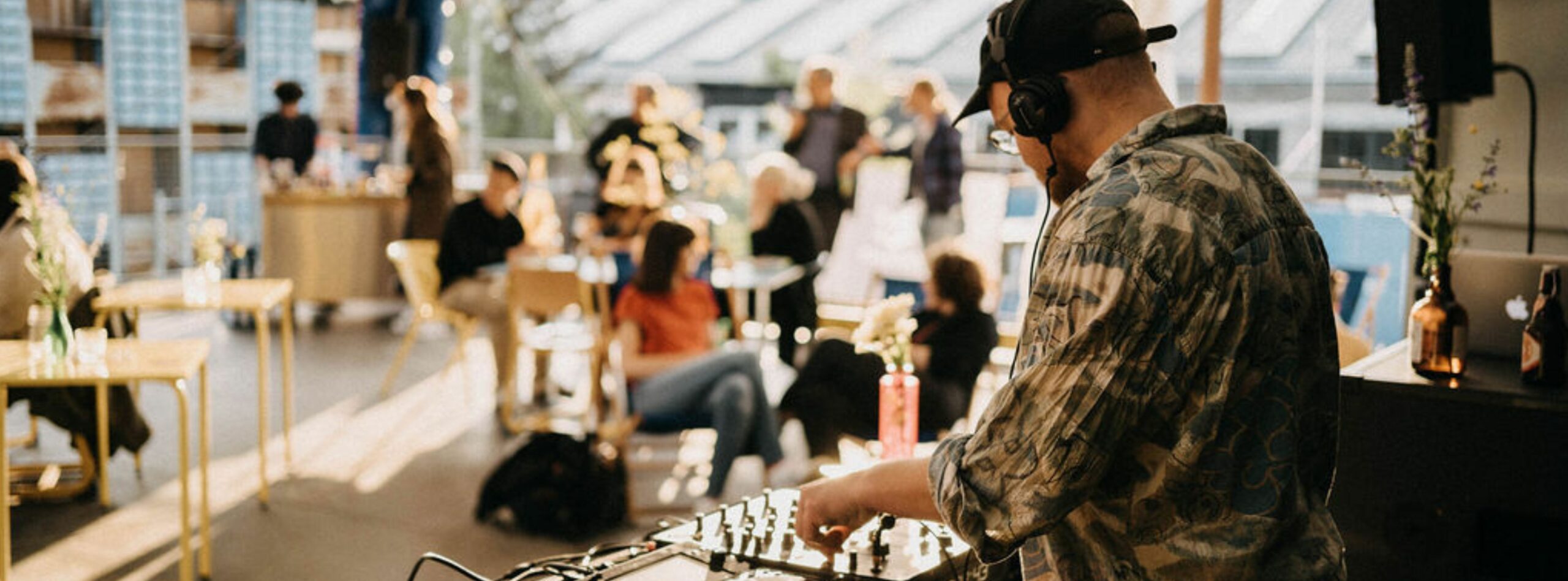 Ein DJ mit Kopfhörern und gemustertem Hemd bedient bei einer Veranstaltung im Freien ein Mischpult. Im Hintergrund treffen sich Menschen in legerer Kleidung, einige unterhalten sich, in einem sonnendurchfluteten Freiluftraum mit Tischen und Dekorationen, alle zusammengeführt durch den Rhythmus der Musik.