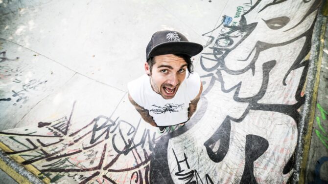 Eine Person mit schwarzer Kappe und weißem T-Shirt steht in einem Skatepark mit Graffiti-bedeckten Rampen und blickt mit offenem Mund in die Kamera. Die Oberfläche des Skateparks ist mit verschiedenen bunten Sprühfarben-Designs und Tags geschmückt.
