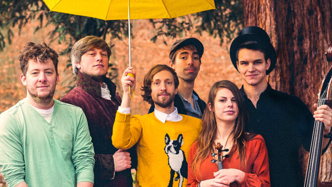 Eine Gruppe von sechs Personen steht draußen in einem Waldstück. Eine Person hält einen gelben Regenschirm und eine andere eine Geige. Sie tragen farbenfrohe, vielseitige Kleidung, darunter einen senfgelben Pullover, ein orangefarbenes Kleid und einen schwarzen Zylinder. Eine Person hält einen Koffer.