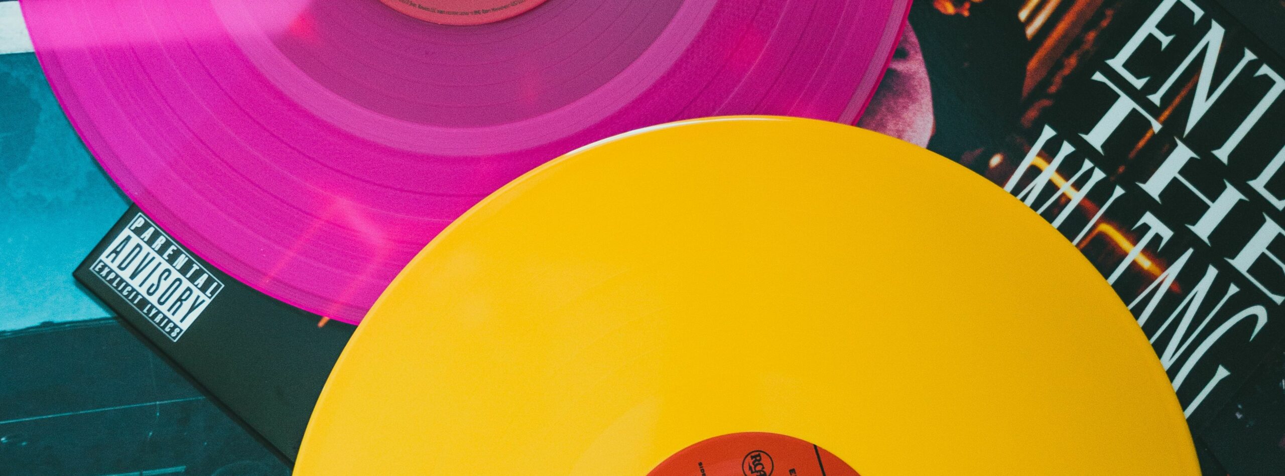 Nahaufnahme von zwei Vinyl-Schallplatten, eine rosa und eine gelbe, die schräg über Albumcovern platziert sind. Die rosa Schallplatte verdeckt teilweise das Etikett mit der Elternberatung auf einem Cover, während die gelbe Schallplatte ein anderes Album mit sichtbarem Text über Pop-Webinare überlappt.